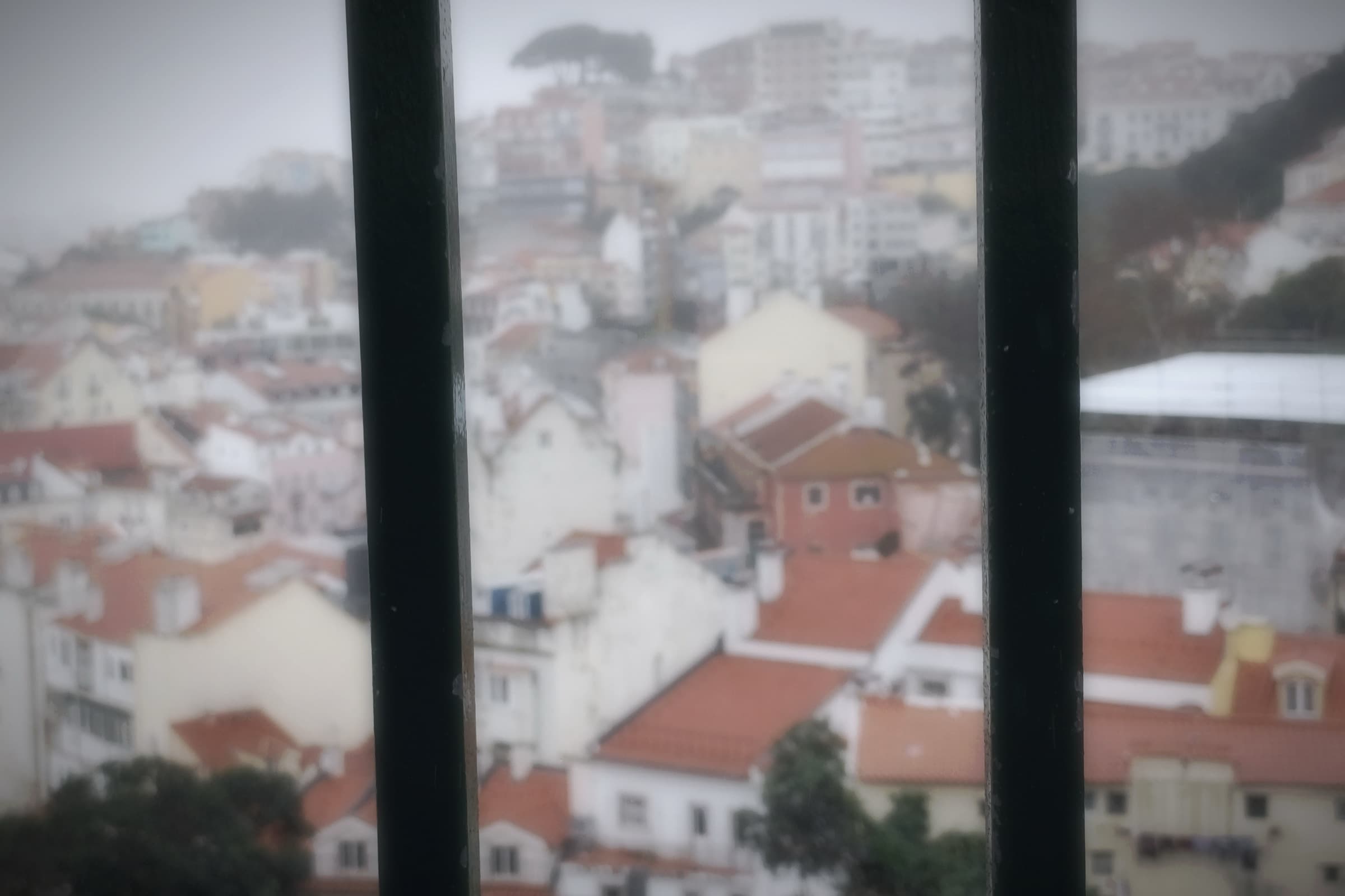 A blurred Lisbon view through dark window bars, with red rooftops softened by fog.