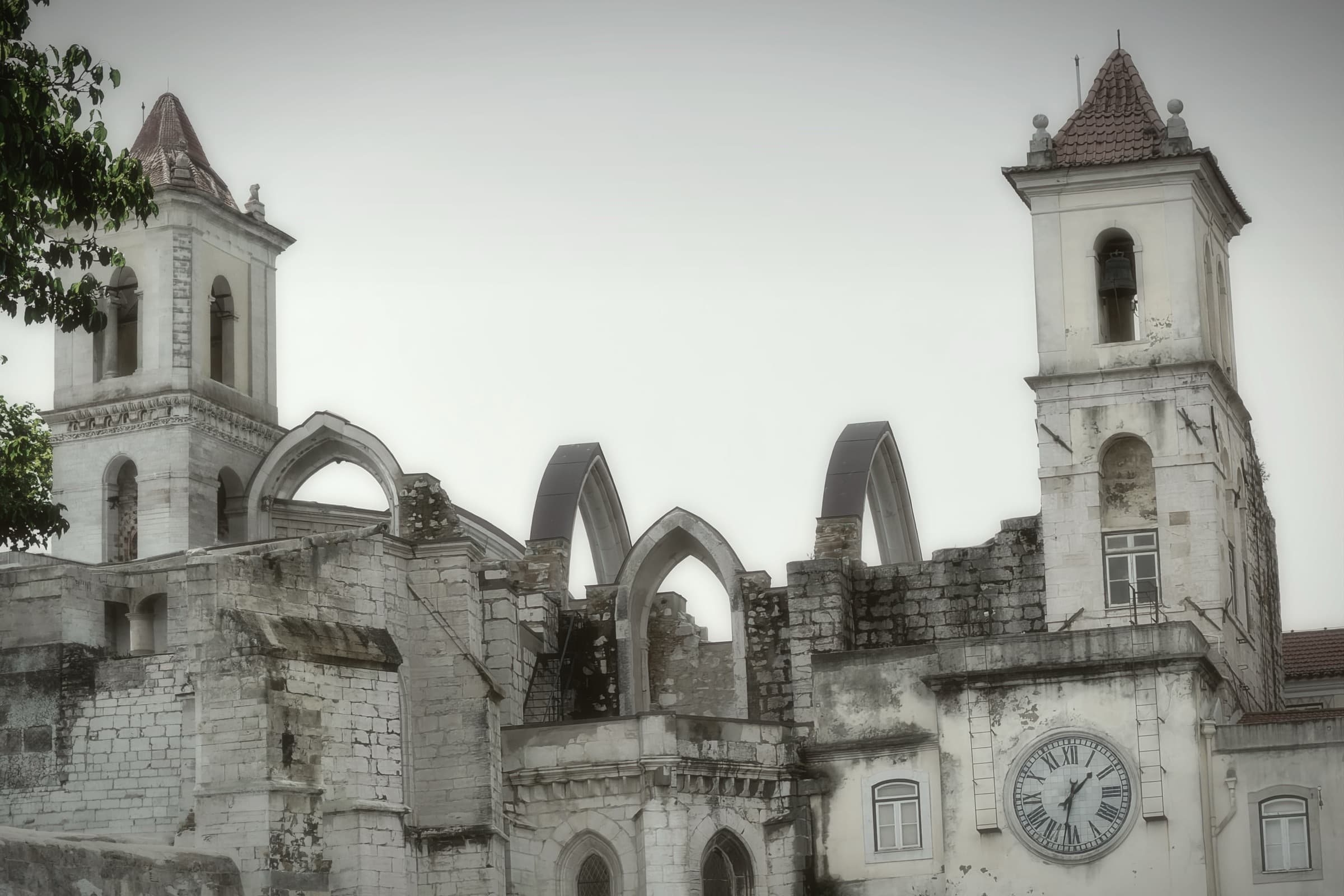 A pale clock tower and ruined stone arches in Lisbon under a soft gray sky.